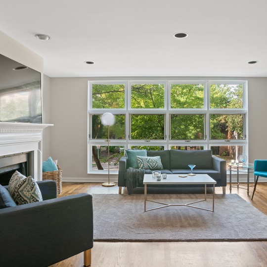 Living room with a fireplace, gray sofas, and large windows overlooking leafy trees.